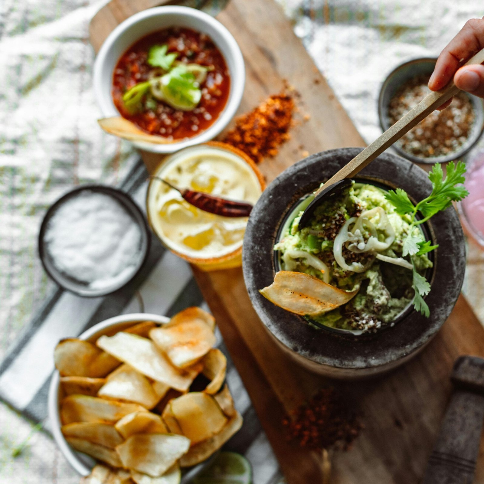 Hand preparing guacamole with colorful dips, chips, and a recipe book on a wooden board.