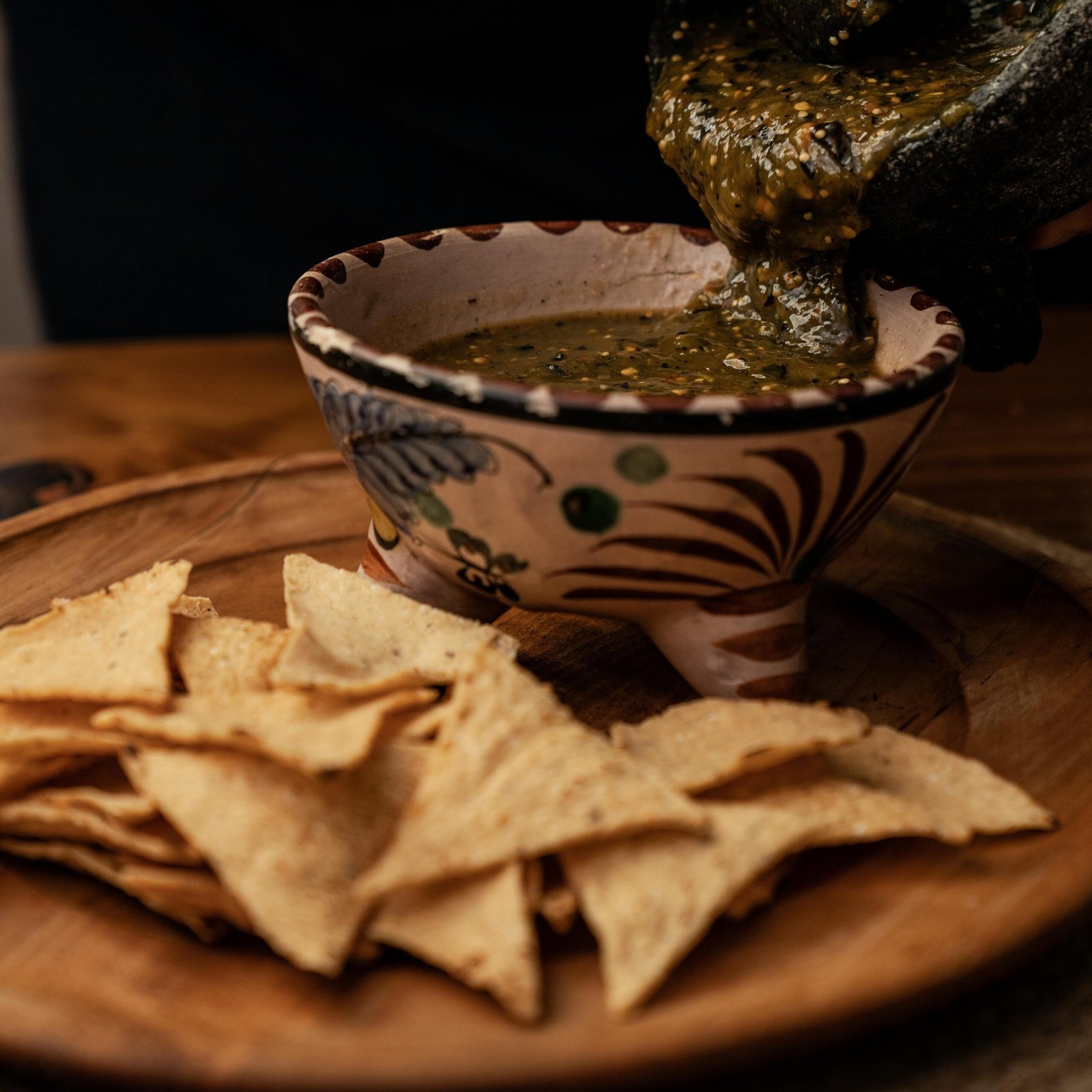 Hands preparing salsa in a traditional mortar, served with nachos on a wooden plate.